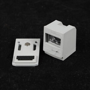 May include: A light gray miniature computer model with a separate keyboard component. The computer has a screen and a slot for a disk drive. The keyboard has a small black keyboard and two exclamation point shaped holes.