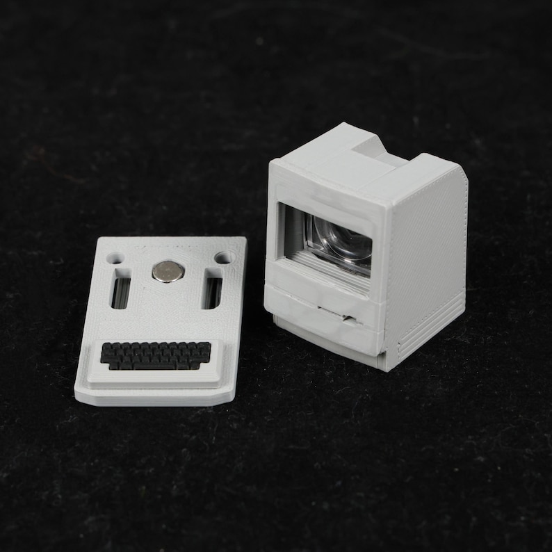 May include: A light gray miniature computer model with a separate keyboard component. The computer has a screen and a slot for a disk drive. The keyboard has a small black keyboard and two exclamation point shaped holes.