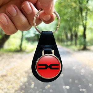 Puede incluir: Un llavero de cuero negro con un llavero plateado. El llavero presenta un emblema circular rojo con un diseño geométrico negro y un borde dorado. El fondo es una escena borrosa al aire libre.