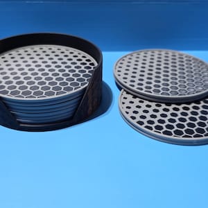 May include: A set of gray and black honeycomb patterned coasters with a black holder. Two coasters are placed separately. The coasters are round and made of a durable material. The set is on a blue surface.