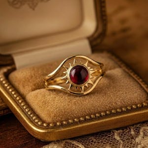 Könnte beinhalten: Ein goldener Ring mit einem zentralen, kreisförmigen, dunkelroten Edelstein, der von einem strahlenförmigen Design mit spitzen Zacken umgeben ist. Die Ringschiene teilt sich in zwei geschwungene Linien, die das Sonnenmotiv rahmen.