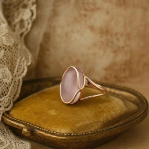 Könnte beinhalten: Roségoldener Ring mit einem großen, ovalen, hellrosa Edelstein. Der Ring hat ein filigranes, offenes Design auf einem goldenen Band. Der Ring wird auf einer goldfarbenen Samtoberfläche präsentiert.