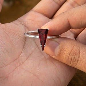 May include: A silver ring featuring a triangular, deep red gemstone. The gemstone is set with small prongs, and the band is a simple, polished silver. The ring is held up in a hand.
