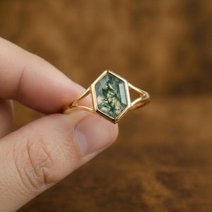 Könnte beinhalten: Ein goldener Ring mit einem sechseckigen Moosachat-Edelstein. Der grüne Stein hat natürliche Einschlüsse und ist in einem goldenen Band mit offenem Design gefasst. Der Ring wird vor einem verschwommenen braunen Hintergrund gezeigt.