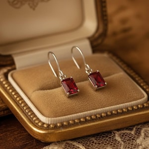 Può includere: Un paio di orecchini pendenti in argento con pietre preziose rettangolari rosso scuro. Gli orecchini sono esposti in un portagioie aperto in stile vintage con una cornice dorata e un interno in velluto beige. Un gioiello classico.