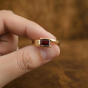 Op de afbeelding: Een gouden ring met een enkele rechthoekige, dieprode edelsteen die horizontaal in een spanningzetting is geplaatst. De ringband is glad en gepolijst en toont een strak, modern ontwerp dat de donkerrode kleur van de steen benadrukt.