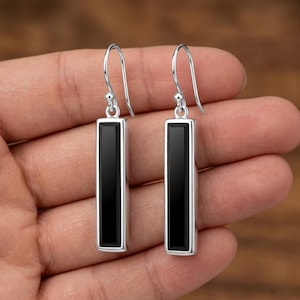 May include: A pair of silver drop earrings with rectangular black onyx stones. The earrings have a simple, modern design with a polished finish. The black stones are set in a silver frame and hang from a curved silver hook.