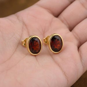 Peut inclure: Deux boucles d'oreilles dorées, chacune ornée d'une pierre précieuse rouge foncé de forme ovale. Les pierres sont serties dans une lunette dorée, créant un look classique. Les boucles d'oreilles sont présentées sur une surface claire.