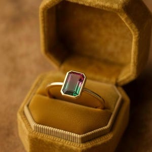 Pode incluir: Um anel de ouro com uma pedra preciosa retangular multicolorida, engastada em uma moldura de ouro. A pedra preciosa transita do rosa para o verde. O anel é exibido em uma caixa de anel de veludo mostarda.