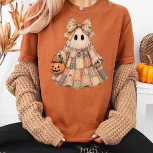 Op de afbeelding: Een roestoranje t-shirt met een afbeelding van een geest in een patchworkjurk met een grote bloemrijke strik. De geest houdt een pompoenemmer vast en draagt een kleine pompoenhanger. Het ontwerp bevat gedempte tinten blauw, beige en bruin.