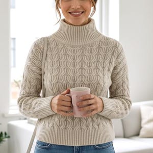 May include: A woman in a beige cable-knit turtleneck jumper and blue jeans, holding a pink mug. The jumper features a textured pattern. A light grey purse hangs from her shoulder. The background includes a window, a sofa, and a potted plant.