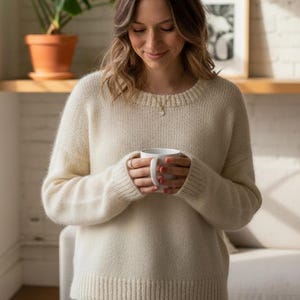 Op de afbeelding: Een persoon in een crèmekleurige trui houdt een witte mok vast. Een Monstera-plant in een terracotta pot staat op een plank. De achtergrond is een witte bakstenen muur en een raam.