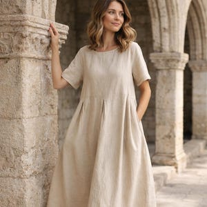 May include: A beige linen midi dress with short sleeves and a pleated bodice. The dress has a relaxed fit and falls to mid-calf. The model is wearing the dress with sandals. The background features stone architecture.