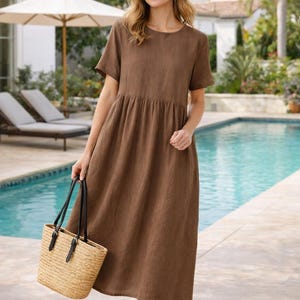 May include: A brown linen dress with short sleeves and a gathered waist. The dress is paired with black sandals and a woven straw tote bag with black leather handles. The background features a swimming pool and outdoor setting.