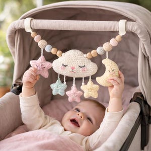 May include: A baby plays with a crochet mobile featuring a cloud, stars, and a moon. The mobile is suspended from a stroller and includes pastel-colored stars and a yellow crescent moon. The cloud has closed eyes and a smiling face.