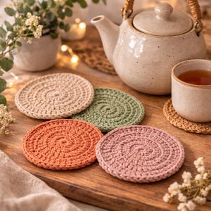 May include: A collection of handmade crochet coasters in various colors, including beige, green, orange, and pink. A ceramic teapot and teacup are in the background, suggesting a cozy tea-time setting. The coasters are arranged on a wooden surface.