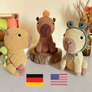 Peut inclure: Trois peluches capybaras crochetées à la main, dans des tons de marron, beige et vert. L'une a une petite citrouille sur la tête, une autre une tortue, et la troisième un chapeau de grenouille. Les peluches sont exposées sur une surface en bois.