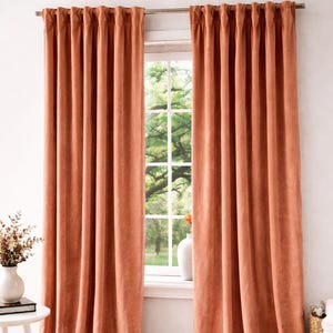 Pode incluir: Cortinas de veludo cor de terracota penduradas em uma barra de prata, emoldurando uma janela com vista para árvores verdes. Uma mesa lateral branca com um vaso de flores e uma cesta de vime estão em primeiro plano. Um tapete bege cobre o chão.