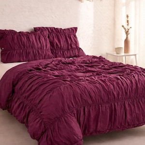 May include: A burgundy bedspread and two matching pillow shams with a ruffled design. The bedding is on a wooden bed frame in a well-lit bedroom. A macrame shelf and a small side table are also visible.