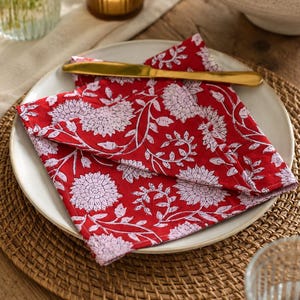 Puede incluir: Una servilleta roja con estampado floral blanco descansa sobre un plato blanco, con un cuchillo dorado. El plato está sobre un salvamanteles tejido. Una vela en un portavelas de cristal y un jarrón con flores están en el fondo.
