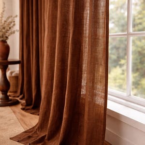 May include: Floor-length brown linen curtains. The curtains have a textured appearance and hang in front of a window. The window has a white frame and looks out onto a green, leafy background.