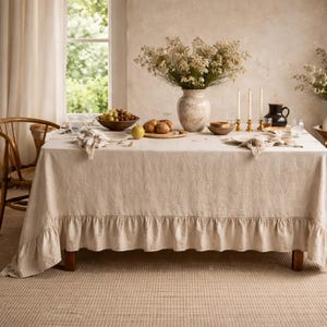 Op de afbeelding: Een beige linnen tafelkleed met een gefranjerde rand bedekt een houten tafel die is gedekt voor een maaltijd. De tafel is gedekt met borden, servetten, kaarsen en een vaas met bloemen. Een kom met druiven en een houten stoel zijn ook zichtbaar.
