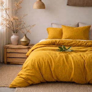 May include: A mustard yellow duvet cover and two matching pillowcases on a bed. The bedding is made of a soft, textured fabric. A wooden crate holds two vases with dried branches. A woven pendant light hangs above the bed.