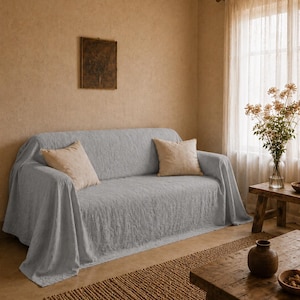 May include: A light gray textured sofa cover draped over a three-seater couch, featuring a subtle crinkled fabric pattern. Two square beige throw pillows are placed on the seat, providing a neutral contrast to the light gray textile covering.
