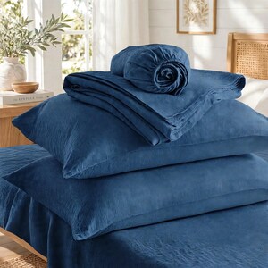 May include: A set of deep blue bedding featuring a textured, crinkled fabric finish. The collection includes a fitted sheet, a flat sheet, and two pillowcases stacked neatly on a bed, showcasing a consistent solid dark blue color throughout.