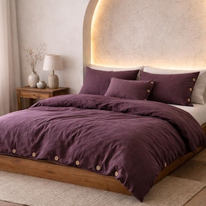 Può includere: Un letto con un copripiumino viola scuro e federe abbinate. Il piumino ha chiusure con bottoni in legno. Il telaio del letto è in legno e la stanza ha una palette di colori neutri con un tappeto beige e un comodino in legno.