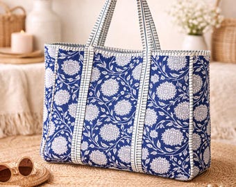 May include: A large tote bag with a blue floral pattern and white accents. The bag has sturdy handles and a rectangular shape. The background includes a straw mat, sunglasses, and a hat.