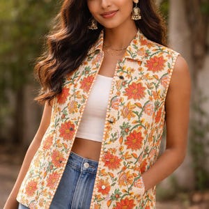 May include: A sleeveless floral print vest in cream, orange, and green. The vest features a button-up front, a collared neckline, and two front pockets. The vest is paired with a white top and blue jeans.