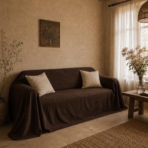 May include: A dark brown sofa cover with a textured fabric finish, draped over a couch. Two square, light beige throw pillows are placed on the seat. The cover extends over the arms and down to the floor, providing a full, loose-fitting drape.