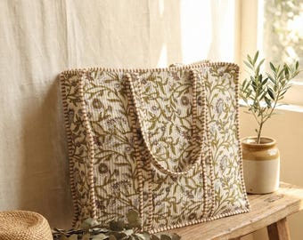 May include: A large tote bag with a floral pattern in green and brown on an off-white background. The bag has a matching handle and trim. A straw hat and eucalyptus branches are on a wooden bench.