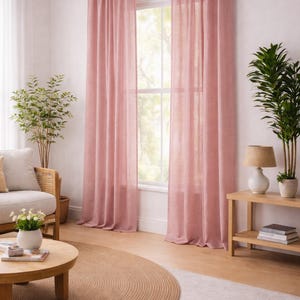 May include: A living room with pink curtains framing a window. A round jute rug sits on a light-colored rug. A wooden coffee table, a sofa, and a side table with a lamp and plants complete the scene.
