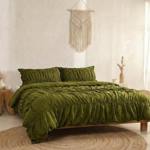 Può includere: Un letto con biancheria da letto verde oliva, tra cui un piumino e due cuscini. Il letto è su una struttura in legno e un tappeto tessuto è sul pavimento. Un arazzo in macramè e vasi decorativi sono anche nella stanza.