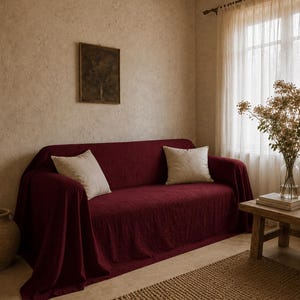 May include: A burgundy sofa draped with a matching cover and two beige throw pillows. The room features a neutral color palette with a woven rug, a small wooden table, and a framed artwork on the wall.