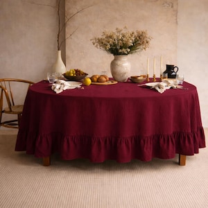 May include: A round burgundy tablecloth with a ruffled edge covers a wooden table. The table is set with plates, glasses, and a vase of flowers. A wooden chair is visible to the left.
