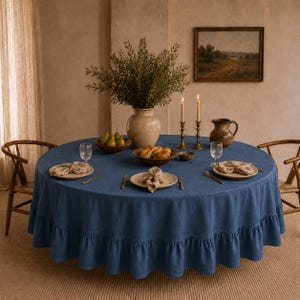 Può includere: Un tavolo da pranzo rotondo, apparecchiato con una tovaglia blu con bordo a balze. Il tavolo è apparecchiato con piatti, posate e tovaglioli. Un vaso con verde, candele e un dipinto completano la scena.