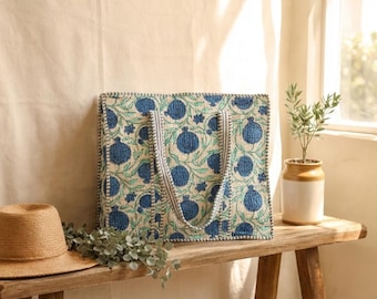May include: A square tote bag with a blue and green floral pattern, featuring a repeating design of stylized pomegranates. The bag has matching handles and a contrasting striped trim. A straw hat and greenery sit on a wooden bench.
