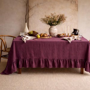Puede incluir: Un mantel rectangular de lino morado oscuro con un borde con volantes, colocado sobre una mesa de madera. La mesa está decorada con un jarrón de flores, velas y platos de fruta y pan, creando una escena de comedor cálida y acogedora.