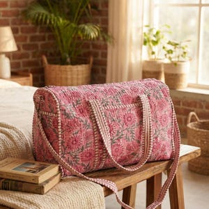 Puede incluir: Bolsa de viaje rosa con estampado floral con asas y bandolera a juego. La bolsa está hecha de tela acolchada y se apoya en un banco de madera. La bolsa tiene forma rectangular y está diseñada para viajar.