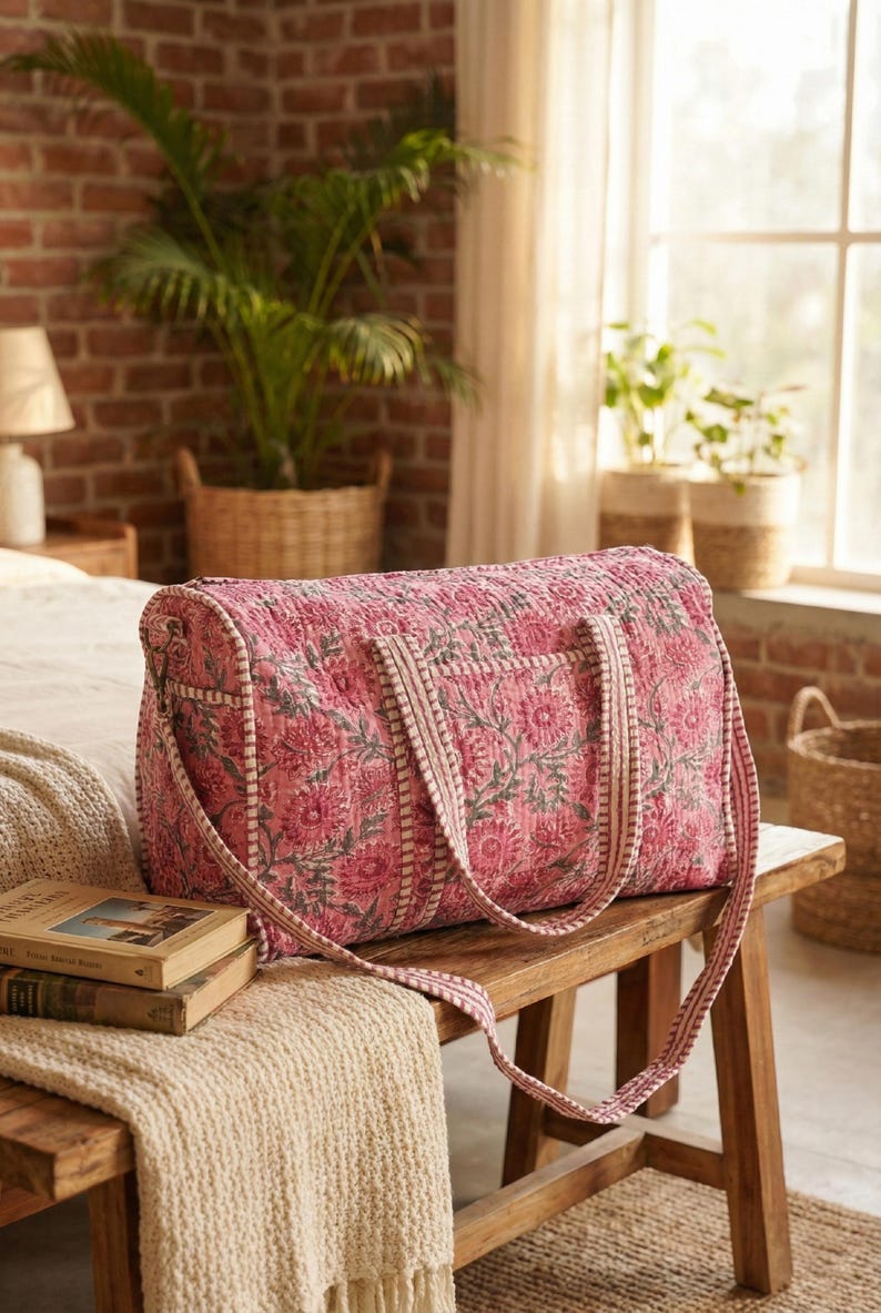 May include: Pink floral print duffel bag with matching handles and a shoulder strap. The bag is made of quilted fabric and sits on a wooden bench. The bag has a rectangular shape and is designed for travel.