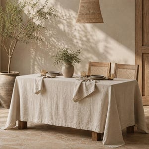 May include: A rectangular dining table covered with a solid beige linen tablecloth, featuring two matching folded napkins placed on the surface. The table is set with ceramic plates and glassware, accompanied by two wooden chairs with cane backrests.