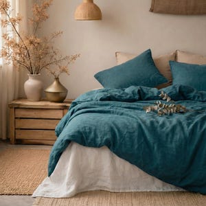 May include: A teal linen duvet cover and pillow set on a bed with a white bed skirt. The bed is in a neutral-toned room with a wooden nightstand, vases, and a woven pendant light. The bedding is arranged with decorative pillows.