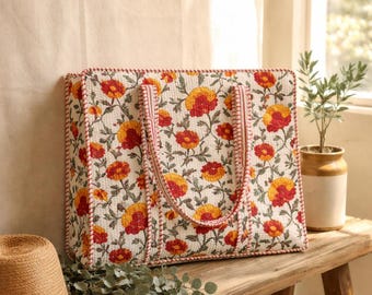 May include: A floral tote bag with a white background and a pattern of red and yellow flowers. The bag has a rectangular shape with a red stitched border and matching handles. A straw hat and greenery are on a wooden bench.