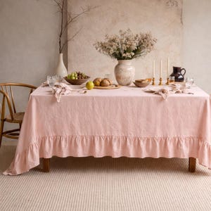 May include: A pink linen tablecloth with a ruffled edge covers a wooden table set for a meal. The table is adorned with a vase of flowers, candles, and place settings. A wooden chair is visible on the left.