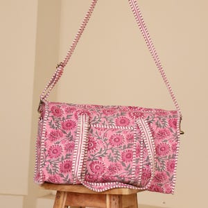 May include: A pink floral print duffel bag with a matching shoulder strap. The bag features a repeating pattern of pink flowers and gray leaves on a pink background. The strap and trim are white with pink stitching.
