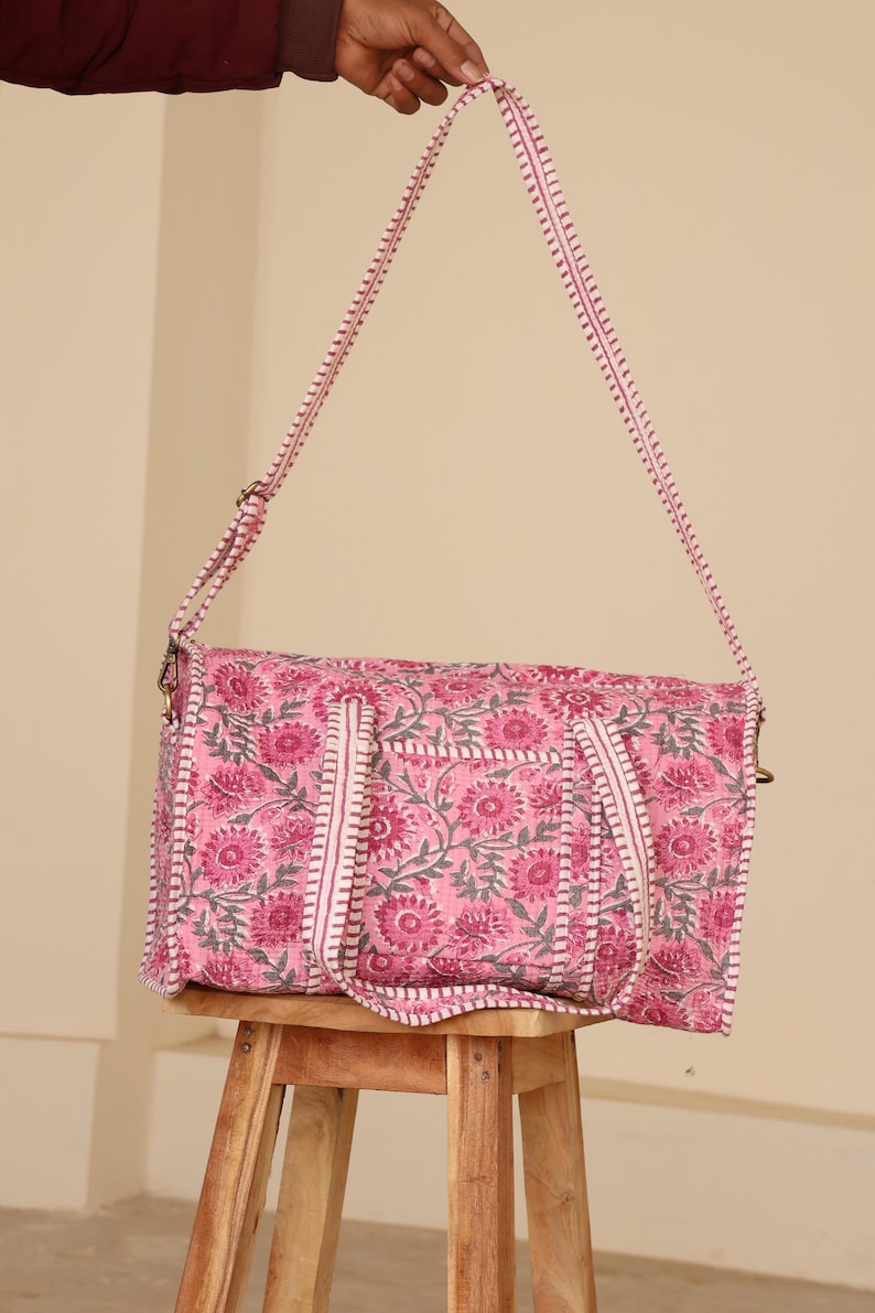 May include: A pink floral print duffel bag with a matching shoulder strap. The bag features a repeating pattern of pink flowers and gray leaves on a pink background. The strap and trim are white with pink stitching.