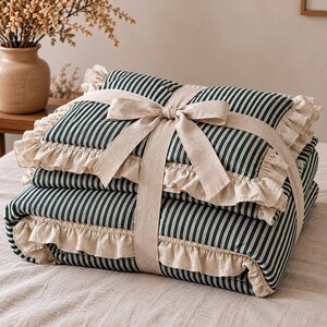 May include: A folded quilt featuring a dark green and white vertical striped pattern. The quilt is edged with a ruffled beige trim and is secured with a wide, matching beige fabric ribbon tied into a bow across the center of the folded layers.